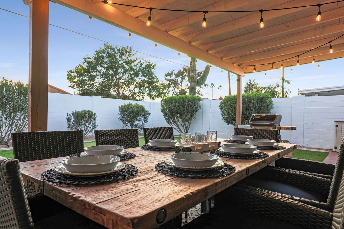 This exterior image features a welcoming patio setup with a wooden table, woven chairs, hanging lights, and an outdoor grill—ideal for alfresco meals and social gatherings amid the Scottsdale climate.