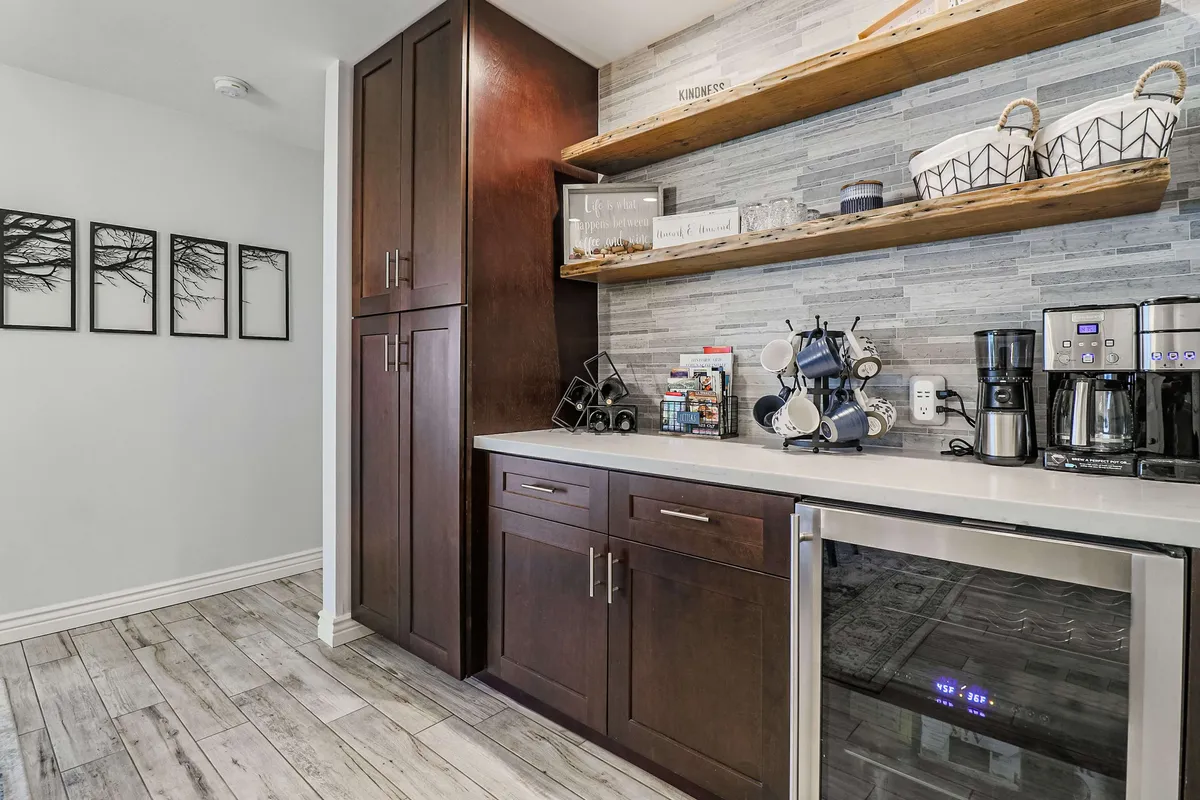A cozy nook within the kitchen features dark wood cabinetry, an open shelf with glassware, and multiple coffee machines, creating a dedicated coffee and wine bar. Artful touches make this spot perfect for socializing or quiet moments.