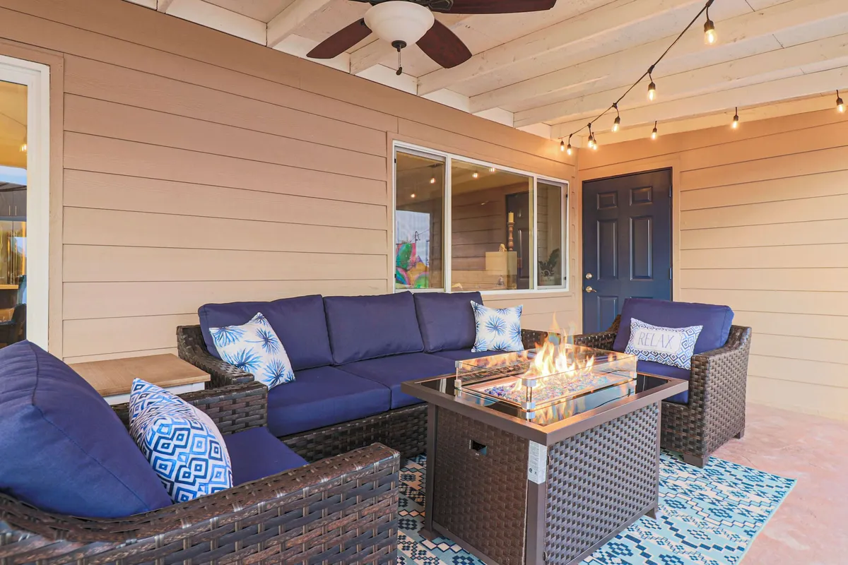 A cozy backyard patio featuring woven seating, a fire table, decorative pillows, and string lights. The setting is ideal for relaxing evenings, socializing with friends, or unwinding after a day exploring the city.